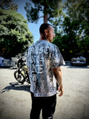 Man wearing a t-shirt outdoors, with a motorcycle and trees in the background.